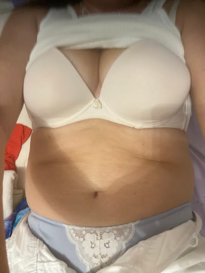 The image shows a close-up of a person lying down, wearing a white bra and light blue underwear with white lace trim. The person's torso is visible, with the belly button clearly showing. The background consists of bedding in shades of purple and white. A small section of red fabric is visible at the bottom of the frame. The lighting suggests an indoor setting, possibly a bedroom. There is no visible text in the image. The overall impression is intimate and casual. The location context of Novi Petrivtsi, Ukraine, does not provide any specific visual cues within the image itself.
