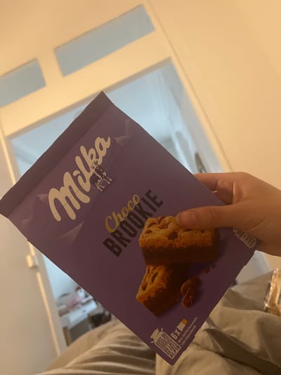 A person's hand holds an open, purple Milka Choco BROOKIE snack package, revealing two individual brookies inside. The brookies appear to have a chocolate chip cookie base topped with a dark chocolate layer. The packaging is prominently branded with the Milka logo and features a cow mascot.
Visible text on the packaging includes "Milka," "Choco BROOKIE," and phrases in both French and Dutch: "AVEC DU CHOCOLAT AU LAIT DU PAYS ALPIN" and "Met melkchocolade van Alpenmelk," both translating to "with alpine milk chocolate." The number "6X" is also visible, indicating multiple servings. Nutritional information is partially visible, showing "1949 kJ / 466 kcal" per 100g and "118 kcal" for a single portion.
The background shows a brightly lit indoor environment, primarily consisting of light-colored walls and blurred reflections, suggesting a home setting. Part of the person's striped clothing is visible on the right side of the frame. The overall impression is a casual moment, possibly preparing to eat the snack. The image was taken in an unknown time of day, with no discernible weather conditions or specific external location cues for Lyon, France, other than the context provided, though the multilingual packaging supports a European context.