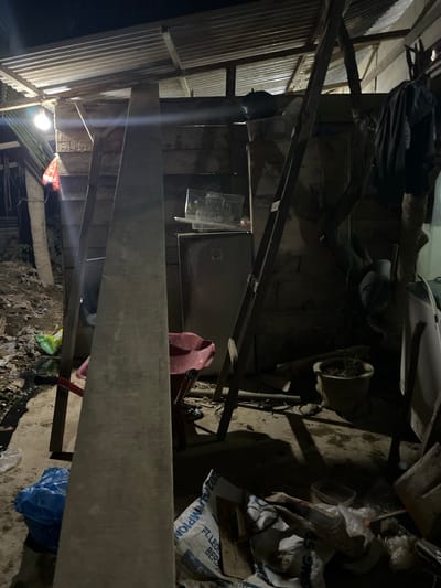 The image depicts a dimly lit, cluttered interior space, likely an outdoor shelter or storage area, located in Benuaraja, Indonesia. A corrugated metal roof is visible overhead, supported by wooden beams. Several objects are scattered around, including a ladder leaning against a wooden wall, a red wheelbarrow, a small refrigerator, and various bags and plastic containers. A bright light bulb illuminates part of the scene. The ground appears to be dirt and debris. The overall impression is one of disarray and a lack of organization.