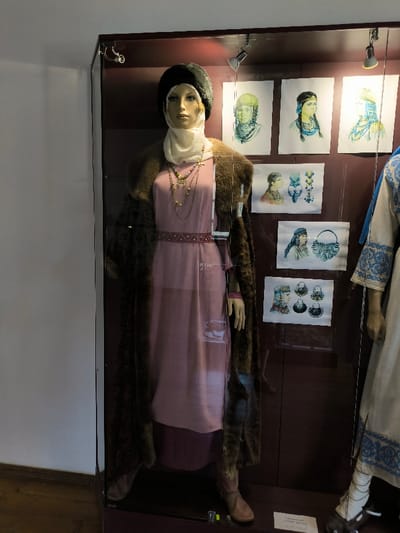 The image displays an indoor museum exhibit located within the Khotyn Fortress complex in Khotyn, Chernivtsi Oblast, Ukraine. A glass display case, illuminated by two internal spotlights, contains two mannequins dressed in historical attire and six mounted illustrations.

The primary subject is a full-height mannequin positioned on the left side of the case. It wears a dark, fur-like headpiece over a cream-colored head wrap. Its face is realistically rendered. The mannequin is clad in a long, light pink gown, cinched at the waist by a narrow, dark red belt featuring a repeating light geometric pattern. A long, dark brown, shaggy coat is draped over its shoulders. Multiple gold-toned necklaces, including various geometric and crescent-shaped pendants and beaded strands, are worn around its neck. Brown, flat-soled footwear is visible on its feet.

To the right, a second mannequin is partially visible. It wears a white garment with blue embroidered patterns, along with a blue fabric draped over its shoulder. White leg coverings and dark, fur-trimmed footwear are also present.

The rear wall of the display case is a dark reddish-brown. Six rectangular illustrations are mounted on this wall. The top row displays three vertical portraits of individuals featuring historical headwear. The middle row contains two horizontal drawings: one depicts a person alongside three individual decorative objects, and the other shows a person next to a decorated bag. The bottom row presents one horizontal drawing of a person with four distinct circular artifacts. The immediate environment surrounding the display case includes a light grey wall to the left and a dark wooden floor section at the base.