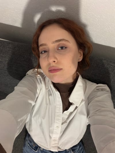 A woman with auburn hair and fair skin lies on a grey couch. She wears a brown shirt underneath a slightly unbuttoned white button-down shirt, with a hint of denim jeans visible. Her face is angled towards the camera, showing her eyes and lips. The background includes a white wall with soft shadows. The image seems to be taken from a slightly high angle, giving a casual, relaxed impression.