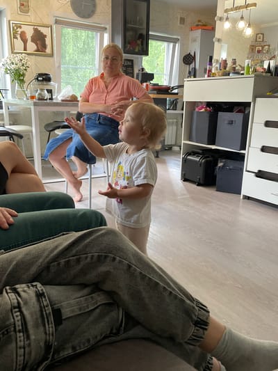 In a kitchen area of a residence in Kumyonki, Russia, a blonde-haired toddler with light skin is standing and gesturing with their hands. They are wearing a white t-shirt with a cartoon graphic. In the background, a blonde woman with glasses and fair skin is seated, wearing an orange button-down shirt and a blue denim skirt. The room features light-colored walls and flooring, a window showing green foliage, and kitchen cabinetry with various items, including stacked black storage boxes, a drawer unit, and a clock on the wall.