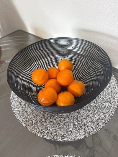 A black wire fruit bowl containing approximately nine tangerines sits on a circular placemat with a black and white woven pattern. The placemat is on a dark, reflective surface, possibly a table. The background consists of a plain, textured off-white wall and a hint of a reflective surface to the left of the wall. The scene appears to be indoors, possibly in a home or kitchen, located in Azalea Park, United States of America. There are no people or animals present. The lighting suggests ambient indoor light.
