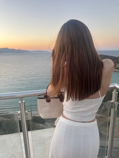 A person, seen from behind with long, dark brown hair, stands on a balcony overlooking the sea at sunset. They are wearing a white, knitted two-piece outfit and carrying a woven straw bag with brown leather accents. The sky displays a gradient of soft oranges and blues, with a distant coastline visible on the horizon. The balcony features a modern railing with glass panels and chrome supports. The scene appears to be in a coastal area, likely a hotel or private residence, with the setting suggesting a peaceful, vacation-like atmosphere. The specific location is the city of Agios Nikolaos Municipal Unit, Greece.