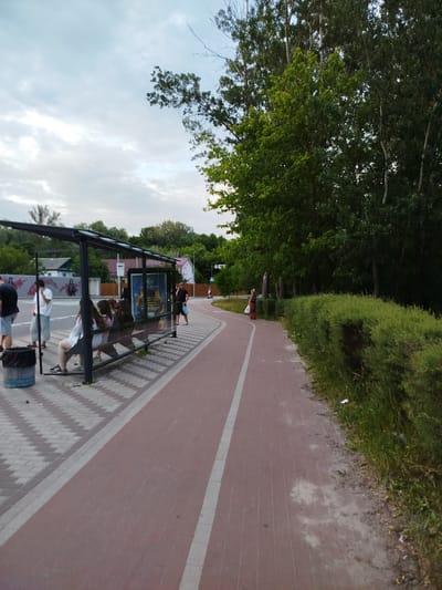 The image shows a bus stop in Chernihiv, Ukraine. Several people are waiting at the bus stop, which has a modern glass and metal shelter. The ground around the bus stop is paved with interlocking bricks, and a red asphalt path, likely a bike lane or pedestrian walkway, runs alongside it. Lush green trees and bushes line the right side of the path. The sky is cloudy, suggesting it's daytime, possibly late afternoon or early evening.
In the foreground, a trash can is visible near the bus stop. People are seated on the bench inside the shelter, while others stand nearby. On the red asphalt path, a woman in a red outfit is walking away from the camera. Further down the path, a person on a bicycle or scooter is visible in the distance, and another woman is walking in the far distance.
A poster is displayed inside the bus stop shelter, featuring text and images, though the details are not clearly legible. The overall scene depicts a typical urban or suburban public transport waiting area with people going about their day. The presence of trees and greenery suggests the area is somewhat suburban or a park-like setting. The weather appears overcast but not actively raining.