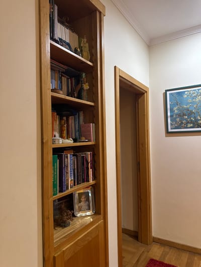 This image depicts an interior residential space within the city of Mirandela, Portugal. The scene is dominated by a built-in, light-stained wooden bookcase on the left, spanning from floor to ceiling. The bookcase contains six open shelves, each filled with various books of different sizes and colors, alongside an assortment of decorative items. These items include a small classical-style statue, several small vases, an Eiffel Tower figurine, a white lidded container, and a small dark spherical object. On the bottommost visible shelf, a silver-framed photograph of a young child is displayed, positioned next to a small dried plant or coral-like ornament.
To the immediate right of the bookcase, an open doorway, framed with matching light-stained wood, leads into an unseen room or corridor. The walls throughout the visible area are painted a uniform light cream or off-white. White crown molding is present along the ceiling line. On the wall to the right of the doorway, a framed picture depicting a blossoming tree against a blue sky, reminiscent of "Almond Blossoms," is mounted. The floor features light-toned wooden parquet flooring, with a portion of a red textured rug visible in the lower right corner of the frame. Artificial lighting illuminates the area, casting a visible reflection on the glass of the framed photograph. No individuals are present in the scene.