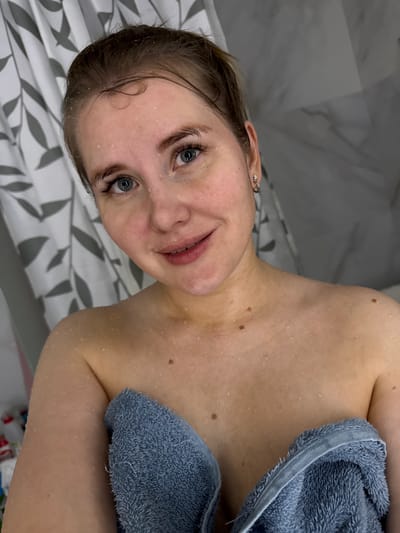 A young, fair-skinned woman with light blue eyes and wet hair is shown in a close-up, angled shot. She is partially covered by a blue towel, and water droplets are visible on her skin. The background features a gray and white patterned shower curtain and a tiled wall. The image appears to be taken indoors, possibly in a bathroom. There's no indication of any location or relation to a city, Russia.