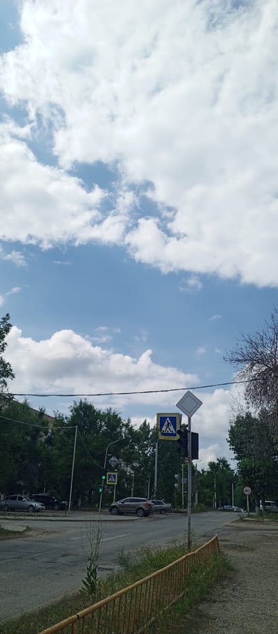 The image depicts a street scene in Zhirnovsk, Russia, on a cloudy day. In the foreground, a weathered yellow metal fence runs along a grassy embankment. The road, which appears somewhat worn with visible cracks, curves to the right. A traffic light with a green signal is visible, accompanied by a blue pedestrian crossing sign featuring a white silhouette of a person walking. Further along the road, another pedestrian crossing sign is mounted higher on a pole, along with a white diamond-shaped sign. A speed limit sign indicating "40" is present on the right side of the road. Several cars are parked and driving on the road, including a silver sedan, a dark SUV, and other vehicles further in the distance. Tall green trees line the sides of the road, with some appearing to be deciduous. The sky above is filled with a mix of blue sky and large, white, puffy clouds.