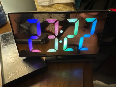 The image displays a digital alarm clock with LED numbers showing the time. The numbers are illuminated in a gradient of colors, transitioning from blue on the left to pink in the middle and green on the right, with the colon between the hours and minutes also displaying a gradient. The clock face is reflecting a dimly lit room, with portions of a person's upper body and clothing visible in the reflection. A metallic object, possibly a power bank with "BEEPOW" embossed on it, is situated to the left of the clock. The clock and the metallic object are placed on a wooden surface, likely a table or desk. The background is out of focus and appears to be a domestic setting. There is no discernible information in the image that would indicate a specific location in Małkowo, Poland.