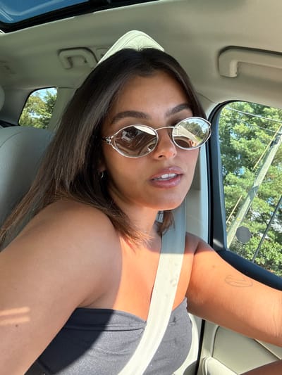 A female individual is seated in the driver's position of a vehicle in West Windsor, United States, taking a selfie. She has dark brown hair, dark eyebrows, and is wearing silver-framed, oval sunglasses with reflective lenses, a dark strapless top, and a light-colored seatbelt across her chest. Direct sunlight illuminates her right side, revealing tanned skin and a faint, linear tattoo on her right upper arm. Her lips are slightly parted. The vehicle's interior features a light-colored headliner, a visible sunroof, and an overhead grab handle. Through the driver's side window and reflected in her right sunglass lens, green foliage, utility poles, overhead wires, and a road are visible under a clear blue sky. The reflection in her left sunglass lens shows architectural elements, possibly from a nearby building.
