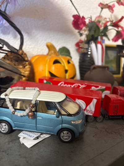 The scene, located in the city of Kuklen, Bulgaria, depicts an indoor arrangement of decorative items and toys on a dark, textured surface. In the foreground, a light blue toy minivan with a white roof is positioned, featuring a white beaded bracelet with a small copper-toned bell charm draped over its front roof and windshield. Beneath the driver's side of the minivan, a crumpled white paper document or tag is partially visible. Immediately behind the minivan, two red toy semi-trailer boxes, bearing the Coca-Cola logo and images of a bottle and holiday ornaments, are arranged. Further into the midground, a carved orange Jack-o'-lantern pumpkin is visible. The background is out of focus, revealing a dark, ornate structural element on the left, and on the right, a dark vase containing several pink and red artificial flowers, behind which a rounded dark brown ceramic vessel and a framed item are discernible. The lighting suggests an ambient indoor environment.