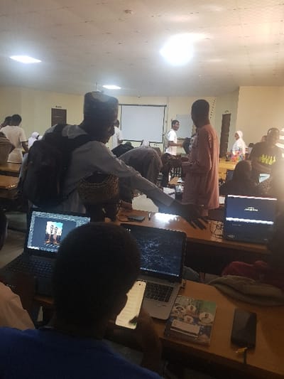 The image captures an indoor scene within a classroom or workshop in Panhauya, Nigeria. Approximately 15 to 20 individuals, predominantly young adults, are present. The room features light-colored walls, a ceiling equipped with multiple square fluorescent light fixtures, and a prominent, bright circular overhead light source. A white projection screen is visible on the rear wall, to the left of which stands a dark brown door.
In the immediate foreground, viewed from behind, a person in a blue shirt is engaged with a smartphone held in their hands. On the wooden desk before them, two open laptops are visible. The laptop on the left displays image editing software (Adobe Photoshop) featuring an image of two stylized figures. The laptop on the right shows a dark desktop background. A book with a multi-face cover and a dark electronic device with a yellow cable are also positioned on the desk.
In the mid-ground, a male wearing a light-colored long-sleeved shirt, a dark kufi-style cap, and a dark backpack leans forward, extending his right arm horizontally. To his right, another male, dressed in a light brown long-sleeved garment, stands facing left. In the background, multiple individuals are observed, including a male in a white t-shirt near the projection screen and a female wearing a white hijab. The scene indicates a collaborative or instructional session, with several individuals actively using laptops.