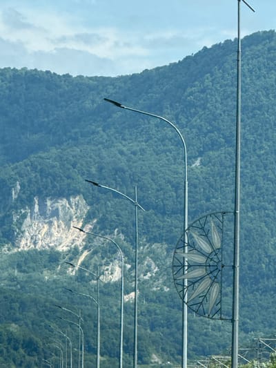 The image shows a mountainous landscape with a row of streetlights leading the eye towards the distance. The mountains are covered in dense green forest, with a lighter, rocky section visible on the left side. The sky is partly cloudy. On the right side of the image, a tall streetlight pole features a decorative, semi-circular metal ornament with a floral or leaf-like pattern. In the far background, what appears to be a power line or electrical infrastructure is visible. The scene appears to be a road or highway in a natural setting. Based on the provided context, this scene is located in Adlerskiy, Russia.
