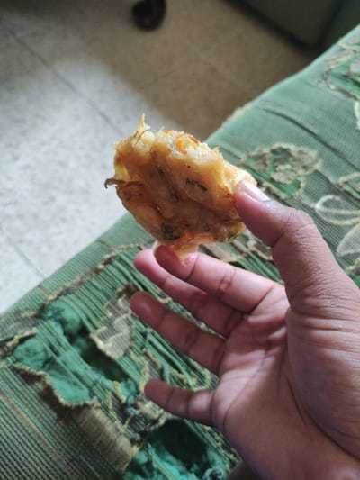 The image shows a hand holding a piece of fried food, possibly a fritter or a pastry. The hand is holding the food up against a green fabric surface, which has a pattern of stripes and is torn in places. The food is crispy and brown, and it appears to have a filling of some kind. The scene is most likely a domestic one, with a sofa or similar furniture in the background. The coordinates provided, -7.828 latitude and 112.701 longitude, place the location in Kecamatan Lawang, Indonesia. There is no additional information available to provide further context about the scene.