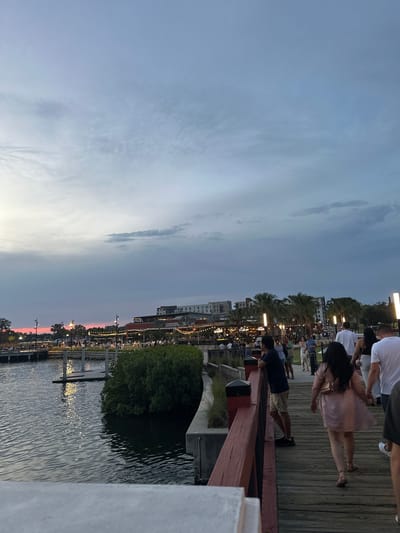 The image shows a waterfront scene in Tampa, Florida, at dusk. A group of people is gathered on a wooden boardwalk overlooking the water. Some people are standing near a red railing, looking out at the view, while others are walking along the boardwalk. Lights illuminate the buildings and the waterfront area. The sky is a mix of blue and gray with hints of pink from the setting sun. The water reflects the lights, creating shimmering patterns. A small green area with trees is visible adjacent to the water.