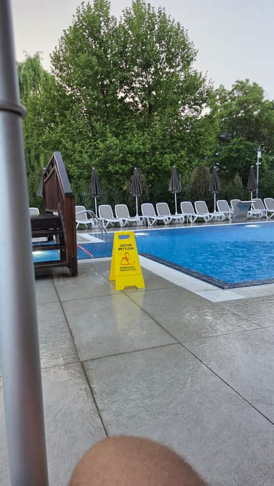 The image shows a wet floor caution sign placed on the tiled area next to a swimming pool. The sign is yellow and triangular, with text in English and what appears to be Cyrillic script. The English text reads "CAUTION WET FLOOR". The swimming pool is filled with blue water and there are ripples on the surface, indicating it may be raining or recently rained. Several empty lounge chairs are arranged around the pool, along with dark umbrellas. Trees and foliage are visible in the background, suggesting an outdoor setting, likely a resort or hotel. A wooden staircase with railings is to the left of the frame, leading down towards the pool. A portion of a metallic pole is also visible in the foreground on the left. The scene suggests a damp, possibly overcast day at a poolside location. The location is identified as Bania, Bulgaria.