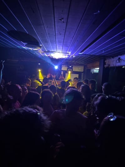 A concert scene unfolds in a dark, intimate venue. Attendees are packed closely together, their faces illuminated by stage lights. Beams of light in blue and yellow colors streak across the scene. A wall with wooden planks is lit with blue light. A doorway can be seen in the background.