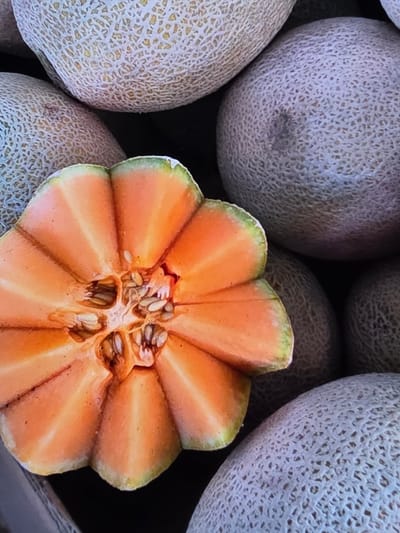 An overhead shot shows a close-up of a halved cantaloupe surrounded by whole cantaloupes. The cantaloupe flesh is a vibrant orange, with visible seeds and stringy pulp in the center cavity. The exterior of the fruit is characterized by its ribbed pattern and a light green rind. The surrounding cantaloupes are mostly visible in their entirety, exhibiting a similar netted rind texture. The image appears to be taken in a market or produce stand setting. No people or animals are present in the frame. The overall impression is one of fresh produce. The image was taken in Voluntari, Romania.