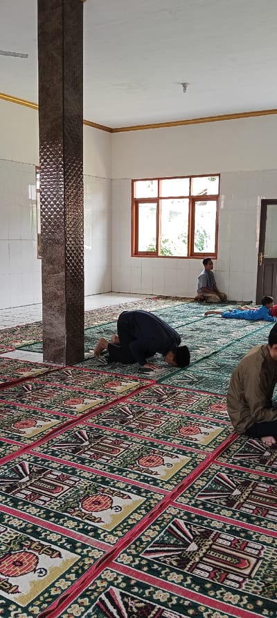 The image shows the interior of a mosque in Kecamatan Kutawaringin, Indonesia (latitude: -6.973, longitude: 107.521). The mosque is decorated with tiled walls and a patterned carpet. There are four men praying on the carpet, all facing the same direction. One of the men is standing, while the other three are kneeling and prostrating themselves in prayer. The mosque has a large window on one side of the room, and there is a door on the opposite side. The door is closed. There is a pillar near the center of the room.  The men are wearing traditional Muslim clothing. There is a light fixture on the ceiling. The room has a white ceiling and white walls. The floor is covered with a patterned carpet.  The carpet is green with red and gold details. The men are facing a wall in the distance. The men are all focused on their prayers.