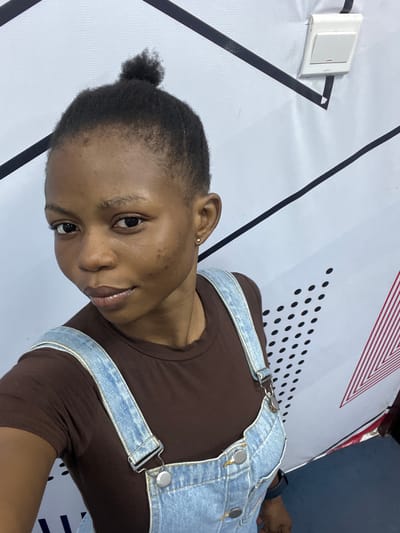 A young woman with short, dark hair pulled back into a small bun is posing for a close-up shot. She is wearing a brown t-shirt and denim overalls. The background features a white surface with black geometric patterns, red stripes, and black dots. A light switch is visible in the upper left corner. The location is Ijanikin, Nigeria, based on available location information.