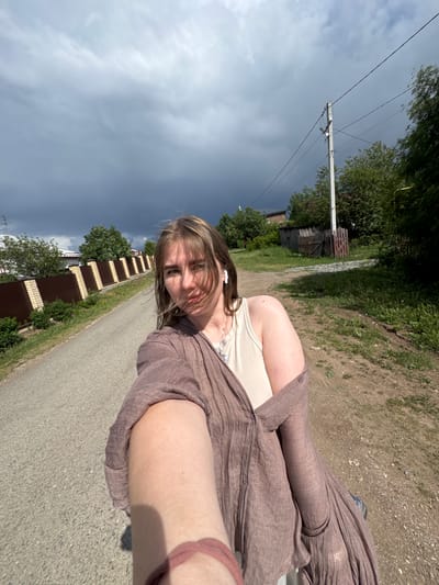 A young woman is outdoors in Горный, Russia, on a paved road between residential areas. She is wearing a brown, button-down shirt over a beige tank top, and earbuds. A fence runs along the road behind her. The sky is overcast with dark clouds. Power lines are visible in the distance.