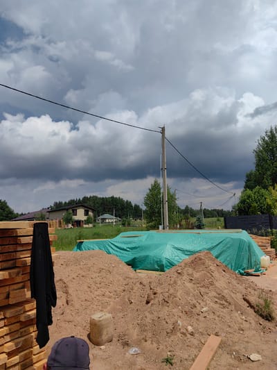 The image shows a construction site with a pile of lumber on the left, a large mound of dirt in the center, and a building covered with a green tarp in the background. There are houses and trees in the distance. The sky is cloudy. A black piece of clothing is hanging on the lumber, and a black baseball cap is in the foreground. A yellow plastic container is sitting on the dirt mound. The location appears to be in a rural area, possibly the village of Гаранскі сельскі Савет, Belarus, given the architectural style of the houses and the surrounding landscape. The overall scene suggests an ongoing construction project in a suburban or rural setting.