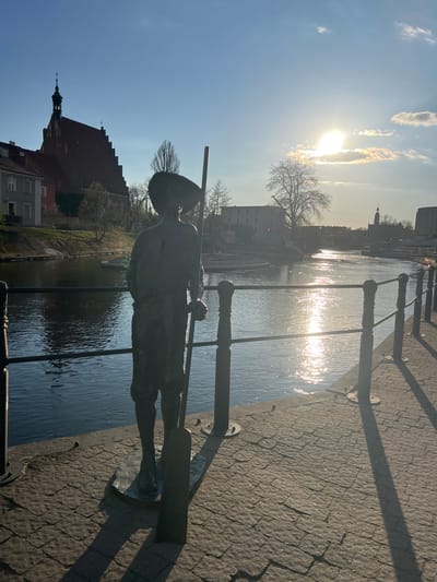 A statue of a young boy stands on a cobblestone walkway beside the Brda River in Bydgoszcz, Poland. The statue depicts the boy with a brimmed hat, carrying a pole across his shoulders, appearing to be in mid-stride. A metal railing runs along the walkway beside the river. The river reflects the sunlight, creating a glare. In the background, various buildings and trees are visible. The sun is high in the sky, partially obscured by clouds.