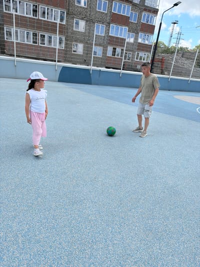 An Asian man and a young girl are on a blue, speckled outdoor court with a chain-link fence in the background. The man, wearing a light olive-green t-shirt and light gray shorts, stands with his arms slightly bent. A green soccer ball is at his feet. The girl, facing left and wearing a white t-shirt with ruffled sleeves, pink pleated pants, a white cap with a pink brim, and white sandals, stands to the left of the man. The court appears to be a sports facility, possibly in Novorossiysk, Russia, given the architecture and utility pole visible behind the fence. The building behind them is under construction, with exposed brick and windows.