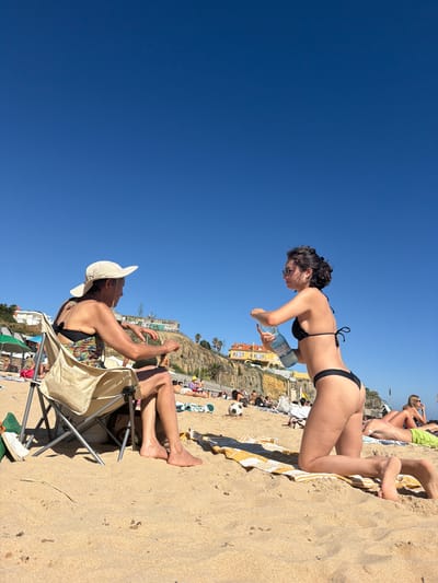 The image depicts a sunny day at a crowded beach in Cascais, Portugal. The sky is a vibrant, clear blue, suggesting fair weather. In the foreground, a man in a white hat and colorful swimsuit sits in a portable beach chair, looking to his right with his hands raised. Beside him, a woman in a black bikini leans forward, holding a large plastic bottle of water. Her posture suggests she is either offering it or preparing to drink.
Further in the background, other beachgoers are visible, some lying on towels, others sitting or standing. The sandy beach slopes gently towards the sea, though the water itself is not directly in view. Behind the beach, a cliff face rises, dotted with buildings and vegetation. The architecture of the buildings, particularly the yellow structure with arched windows, hints at a Mediterranean or Portuguese style.
A soccer ball lies on the sand near a striped towel, indicating recreational activity. The overall atmosphere appears relaxed and summery, with people enjoying the sunshine. The orientation of the image is unusual, with the horizon appearing vertical, suggesting the photo was taken at an angle or that the original camera was tilted. No distinct text is clearly legible in the image, although there might be faint lettering on some of the distant buildings or objects. The time of day is clearly daytime, with strong sunlight casting shadows, indicative of a mid-morning to late afternoon period.