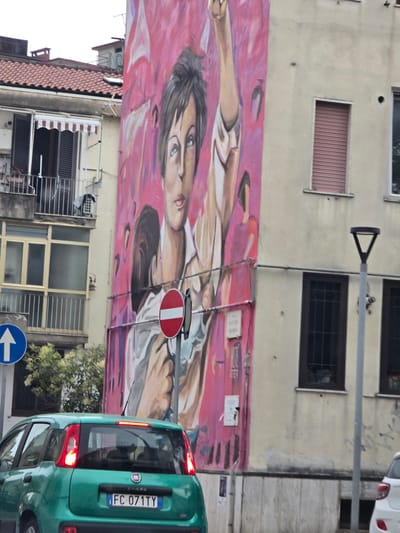 This is a photograph taken on a cloudy day, featuring a large mural on the side of a building in Salerno, Italy. The mural depicts a stylized portrait of a person with a raised arm. In the foreground, a green Fiat Panda car with its brake lights illuminated is visible. The car's license plate reads "FC 071TY". To the left of the car, a blue traffic sign with a white arrow pointing upwards is mounted on a pole. Adjacent to the mural, a street lamp with a triangular top stands on a gray pole. The building's facade is a light beige color, with several windows visible, some with dark shutters. To the left of the mural, a portion of an adjacent building with a tiled roof and balconies is visible. The environment suggests an urban setting with street-level activity.