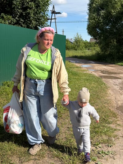 In Andreyevo, Russia, on a rural road with puddles of water, a woman and a young child are walking. The woman, with blonde hair and wearing a pink headband, a light-colored jacket over a green t-shirt with "GEFORCE" printed on it, and loose-fitting jeans, carries a white plastic bag in her left hand. The child, wearing a grey beanie and a grey tracksuit, holds the woman's right hand as they walk. The environment is characterized by grassy verges, a green fence, trees, and a dirt road. In the background, a wooden utility pole is visible. The scene suggests a casual walk in a semi-rural area.