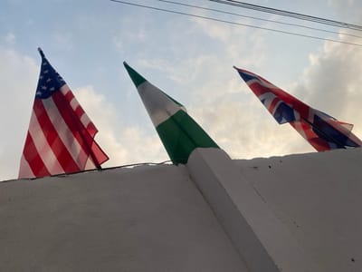 Three flags are displayed atop a white structure against a backdrop of a cloudy sky. From left to right, the flags represent the United States of America, Nigeria, and the United Kingdom. The flags appear to be slightly weathered and angled. A few power lines are visible in the upper portion of the image. The lighting suggests either dawn or dusk.