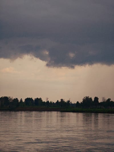 The image shows a wide, calm river with gentle ripples reflecting the subdued light of the sky. The sky is dominated by a large, dark cloud bank in the upper portion, with lighter, patchy clouds below it. Along the far bank of the river, a dense line of dark green trees forms a silhouette against the pale sky. The scene appears to be natural and undeveloped, with no signs of human structures or activity. The location is identified as being in the vicinity of the city of Kazachinskoye, Russia, likely a rural or natural area adjacent to the Yenisei River.