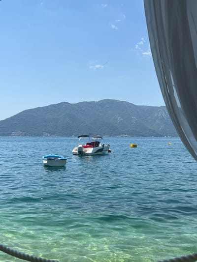 A bright, sunny day is depicted in the bay of Risan, Montenegro. In the foreground, a white sheer curtain hangs on the right side of the frame, partially obscuring the view. The foreground also includes a thick rope. The water is clear and turquoise, with small fish visible beneath the surface. Two boats are anchored in the water. The first boat is a small, light blue and white rowboat. The second boat is a white motorboat with a red canopy and seating. Further in the distance, a large, green, tree-covered mountain rises behind the bay. Scattered yellow buoys are present in the water, and a few distant buildings can be seen on the mountainside. The sky is a clear, bright blue with a few wispy white clouds.
