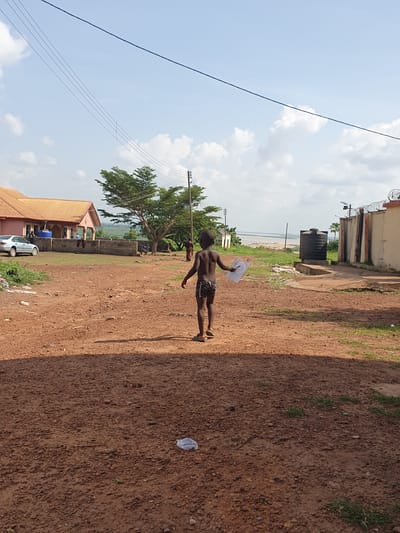 The image depicts an outdoor scene in what appears to be a semi-urban or rural setting in Idah, Nigeria, under bright daylight. The ground is an unpaved, reddish-brown dirt path with scattered patches of green grass. Shadows are visible on the ground, indicating a sunny day.
In the foreground, a young boy is walking or standing on the dirt path. He is shirtless, wearing patterned shorts and flip-flops, and holds a white, thin object, possibly paper or plastic, which has some faint, dark markings or a drawing on it. His back is mostly towards the viewer, and he faces towards the left side of the frame (if rotated correctly).
In the midground, several structures are visible: a building with a terracotta-colored roof and light-colored walls, near which a silver car is parked. Further back, another simpler building has a large, black water tank on its roof, and its perimeter wall is topped with barbed wire. A large, leafy green tree stands prominently, and utility poles with numerous power lines stretch across the sky.
In the distance, beyond the buildings and the tree, a wide body of water, likely a river (consistent with Idah's location on the Benue River), is visible under a hazy sky. The sky above is bright blue with scattered white clouds. The overall scene suggests a typical daily moment in the region. There is no clearly legible text visible in the image.