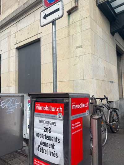An urban street scene in Genève, Switzerland, captured during daytime. The image, rotated 90 degrees clockwise, shows a light-colored, multi-story building facade on the left, constructed with stone or concrete panels. On the right, two newspaper or advertisement stands are visible.
The top stand displays a red banner with "NEWS Immobilier.ch" and the text "Le journal du site n°1 en Suisse romande" (The journal of the No. 1 site in French-speaking Switzerland), along with a QR code and "Plus d'articles. Plus d'annonces. www.immobilier.ch." The lower stand features a similar red banner for "immobilier.ch" and an advertisement for "CHÊNE-BOUGERIES" stating "208 appartements viennent d'être livrés" (208 apartments have just been delivered). The very bottom stand has white graffiti that appears to read "EROR" or "EROF".
In the background, a silver-colored bicycle is leaning against the stands. In the foreground, a metal pole supports a white rectangular traffic sign with a black downward arrow, indicating a one-way street or required direction. Part of a red circular sign, likely a "no entry" or similar prohibition, is also visible next to it. No people are present in the image.