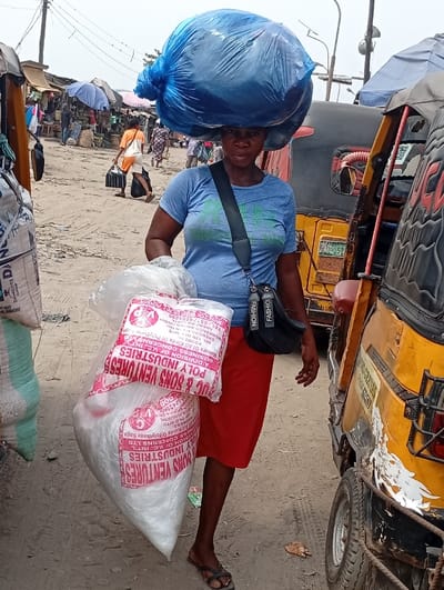 A woman balances a large, blue bag on her head while carrying two large, white bags in her hand. The bags are labeled "BUC & SONS VENTURES POLY INDUSTRIES," with further text in smaller font. The woman wears a blue t-shirt, red skirt, and black sandals. She has a black crossbody bag slung over her shoulder. The background features a bustling outdoor market scene and parked auto rickshaws.