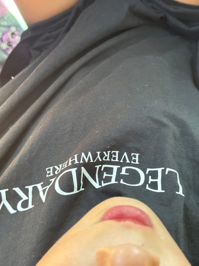 The image shows a close-up of a person wearing a black t-shirt with the word "LEGENDARY" printed in white, vertically, on the front. Below "LEGENDARY" the word "EVERYWHERE" is printed in smaller, white letters. Part of the person's face is visible, including the lips, nose, and cheek. The background is blurry and indistinct, but there is an out-of-focus, colorful object in the lower left corner. There is no evidence indicating a location in Kunje, Montenegro.