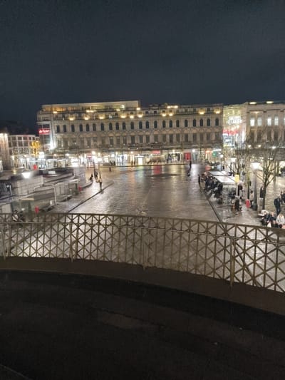The image depicts Place Saint-Lambert in Liège, Belgium, observed from an elevated perspective at night. The sky is dark and featureless.
In the immediate foreground, a metallic railing with a geometric crisscross pattern extends horizontally across the frame, positioned above a dark, indistinct surface.
The midground is dominated by the expansive urban plaza, which is visibly wet and reflective, indicating recent precipitation. On the left side of the plaza, a series of light-toned, angular concrete or stone structures are present, near which several individuals stand or walk. The central area of the plaza is open, traversed by a few solitary pedestrians. On the right side, a covered public shelter or bus stop is visible, populated by approximately 10-15 individuals, some seated on benches and others standing. Bare trees stand adjacent to this shelter and further to the right.
The background features a prominent, multi-story building with a light-colored facade, characterized by multiple arched windows evenly illuminated on its upper levels. The ground floor of this building houses various retail establishments, including a "HEMA" store, with their signs and interiors brightly lit. To the right of this central structure, a large glass building featuring the illuminated sign "GALERIES ST LAMBERT" is visible. Further to the left, another modern building with an "m'aarkt" (or similar) sign and additional illuminated storefronts completes the streetscape. The entire scene is illuminated by artificial streetlights and building lights, with their reflections visible on the wet plaza surface.