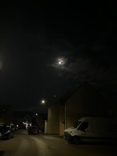 Night scene featuring the moon amidst scattered clouds, accompanied by a slender crescent nearby. A white van is parked along a brick building, with streetlights illuminating the road lined with parked cars. The sky is largely dark, contrasting with the artificial light sources below.