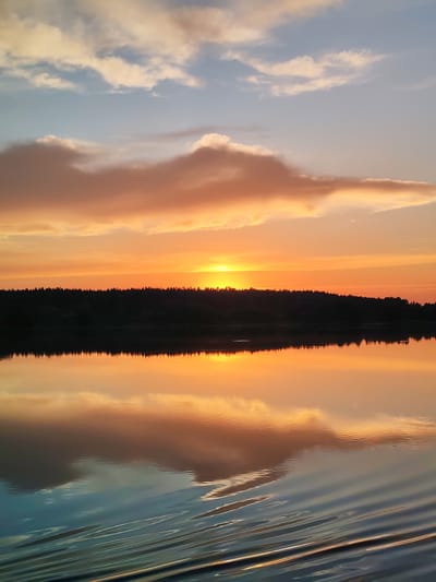 The image depicts a sunset over a body of water. The sky is filled with vibrant orange, yellow, and hints of blue hues from the setting sun, with scattered clouds reflecting the warm light. A silhouetted treeline runs along the horizon, separating the sky from the water. The water itself is calm, creating a clear reflection of the sky and the treeline. In the foreground, subtle ripples disturb the water's surface, creating a textured effect. The location is identified as Terekhovo, Russia. There are no people or discernible objects in the scene other than the natural elements. The overall atmosphere is serene and picturesque.