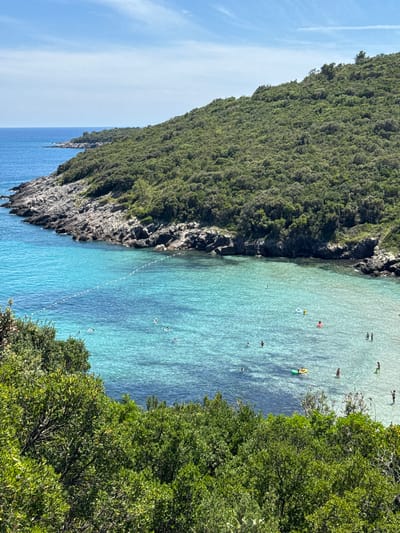 The image depicts a picturesque cove on a sunny day, likely located in the vicinity of Lastva Grbaljska, Montenegro. The scene is dominated by a body of remarkably clear turquoise water that gently laps against a rocky shoreline. The water's transparency allows for a clear view of the sandy seabed. A line of buoys, marked with white floats, separates a section of the bay, likely for swimming or boating.

Several individuals are scattered across the water. Some are swimming, while others are enjoying inflatable water toys, including a yellow ring float and a green raft. A few people appear to be standing in the shallow water near the shore. The cove is framed by a steep, tree-covered hillside that slopes down towards the water. The vegetation is dense and lush, with various shades of green foliage. The sky above is a vibrant blue with a few wispy white clouds. The overall impression is one of a serene and inviting natural landscape, perfect for a summer day by the sea.