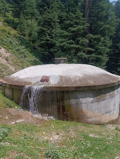 The image shows a large, round concrete water tank situated in a forested area, possibly in Chhyal, India. Water is cascading from a section of the tank's top rim, creating a waterfall effect. The tank has a domed concrete lid with a small metal cap on top and a rusty metal access panel on the side from which the water is flowing. The surrounding environment is a steep, tree-covered hillside. In the background, on the slope, two individuals in casual clothing are visible, walking or hiking. One person is wearing a red backpack. The ground around the tank is covered with grass and some debris, including plastic items. The trees in the background are tall and dense, predominantly evergreens, suggesting a mountainous or hilly terrain. The lighting indicates it is daytime.