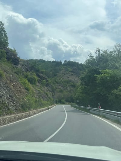 A landscape view, likely taken from inside a moving vehicle on a curving road, presents a mountainous terrain near Rila, Bulgaria. On the right, the asphalt road is marked with a solid white line, bordered by a metal guardrail and vegetation. To the left, a rocky hillside, sparsely covered with greenery, rises steeply. Above the hillside, trees form a line against the sky. Above the trees and hillside is a sky filled with cumulus clouds. The image appears to capture a scenic drive through a mountainous region near Rila.