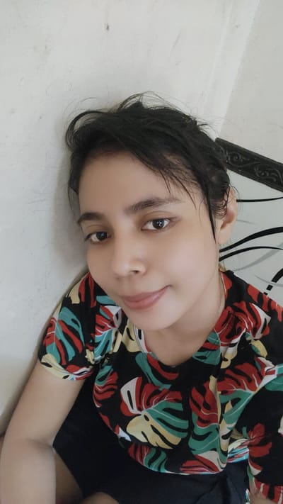 A young woman with dark hair and brown eyes is pictured in a close-up selfie. She is wearing a black t-shirt with a colorful tropical leaf pattern. The background appears to be an interior setting with a textured white wall on the left and a patterned surface on the right, possibly a headboard. The lighting suggests an indoor environment, and there are no visible signs of specific city landmarks or locations within Bekasi, Indonesia.