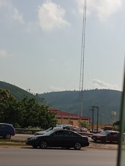 The image depicts a street scene in Kugbo, Nigeria, with a tall communication tower in the background and a hill covered in trees. The sky is pale blue with scattered clouds. In the foreground, several cars are parked along the roadside, including a dark sedan and a blue SUV. Behind the cars is a low building with red roof labeled "INSPECTION CENTRE KUGBO, ABUJA." Utility poles are also visible. In the far background, a water tower can be seen.