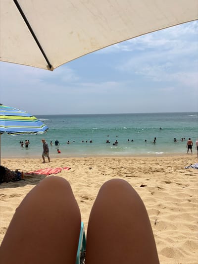The image captures a first-person perspective of a sunny beach day in Puerto Escondido, Mexico. In the immediate foreground, the bare legs of the viewer are visible on the right, suggesting they are seated, possibly on a beach chair, which is partially visible behind the legs. On the left, a large white beach umbrella provides shade, partially obscuring the clear blue sky with sparse white clouds.
The midground features a bustling beach scene. The golden-brown sand, textured with small undulations, stretches towards the turquoise-green ocean water. Numerous people are scattered along the shoreline and in the water. Many individuals are seen swimming, wading, or standing in the shallow ocean, enjoying the clear waters. A man wearing a hat and shorts stands near the water's edge. Further down the beach, a colorful striped beach umbrella, blue and green, adds to the vacation atmosphere. A pink and white striped towel lies on the sand nearby.
The activity depicted is people relaxing and swimming at the beach on what appears to be a bright, clear day, likely late morning or early afternoon, given the sunlight. There is no visible text in the image. The scene conveys a sense of leisure and enjoyment typical of a tropical beach destination.