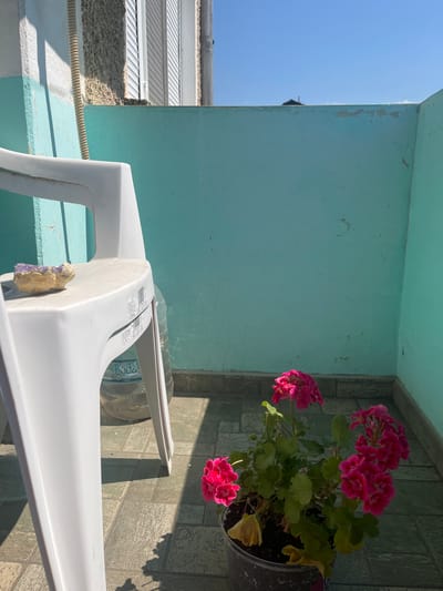 The image depicts a sunny outdoor balcony in Pokrovnik, Bulgaria. A white plastic chair is positioned on the right side of the frame, with a small piece of amethyst resting on its seat. Behind the chair, a large plastic water bottle is visible. The balcony wall is painted a faded teal color and shows signs of cracking. In the lower right corner of the image, a potted plant with vibrant pink flowers sits on the tiled floor, casting a shadow. The sky is clear blue.