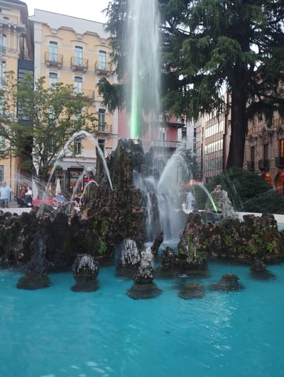 The image shows a fountain with flowing water and rocky structures in the foreground. In the background, there are buildings and trees. On the left, a person in a blue shirt and shorts is standing near the fountain. To the right of the fountain, several people are seated or standing, some near a stroller. The fountain is illuminated with green lights. The scene appears to be in a public area in Lugano, Switzerland.