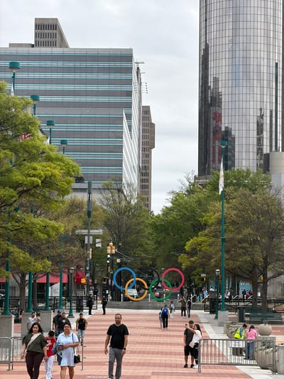 The image shows Centennial Olympic Park in Atlanta, Georgia, United States. In the foreground, pedestrians walk along a brick walkway, some behind metal barricades. In the distance, the Olympic Rings stand, and in the background, several office buildings are visible. Trees line the walkway.